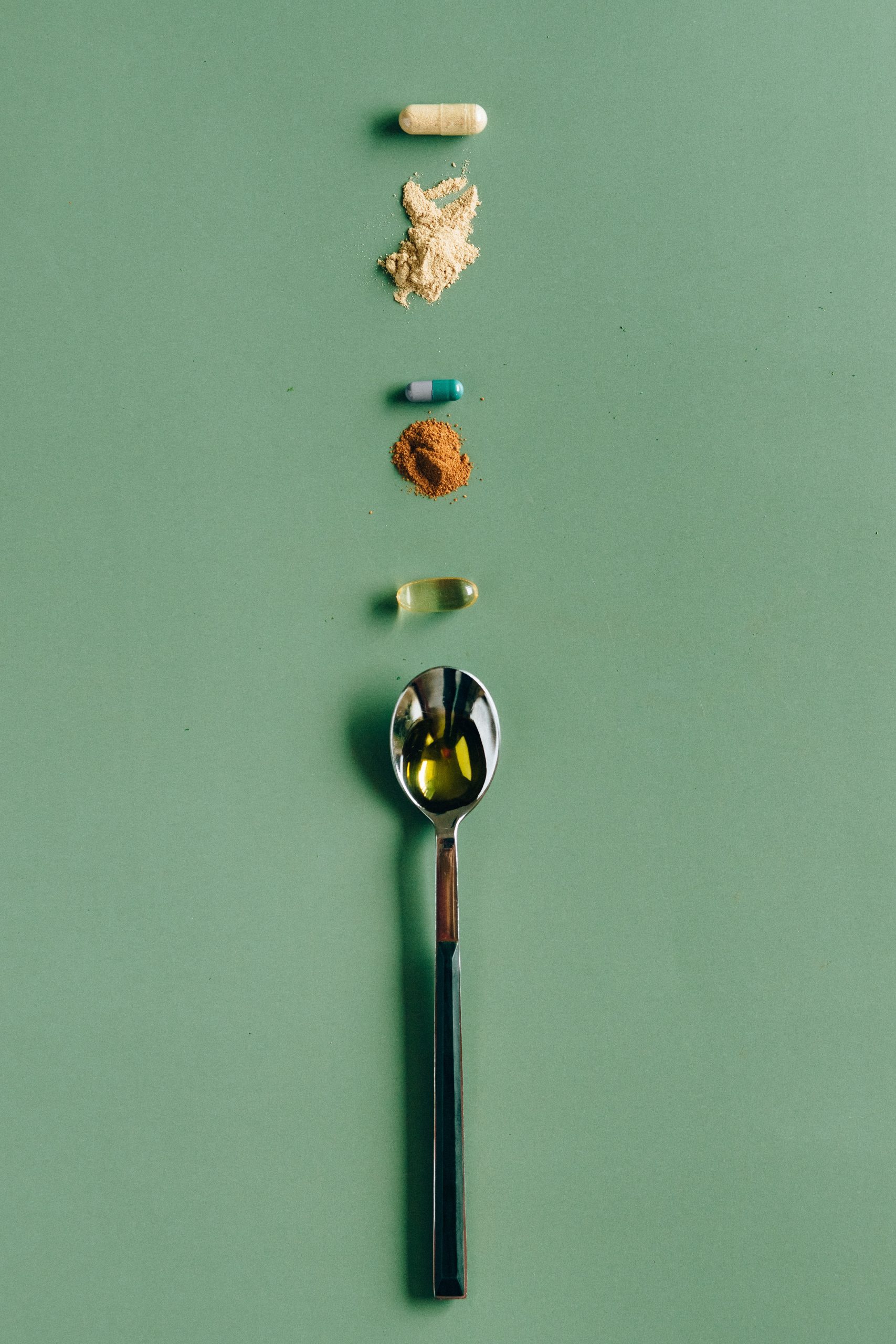 A green background with a silver teaspoon holding liquid. Some pills and tumeric in piles line up with the spoon.